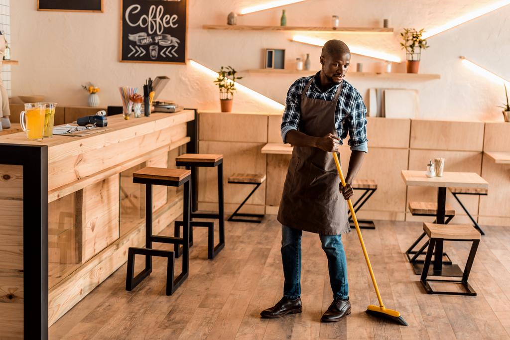african american worker sweeping with broom in coffee shop